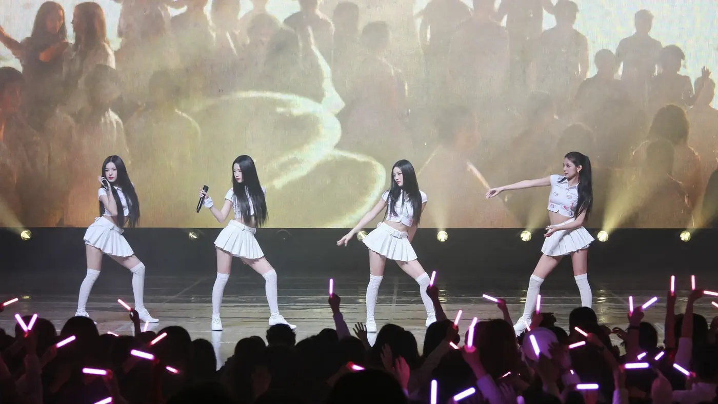 An idol group performing on stage in front of a crowd with light sticks